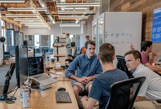 A group of developers talking in a modern office space with laptops and code on the screen